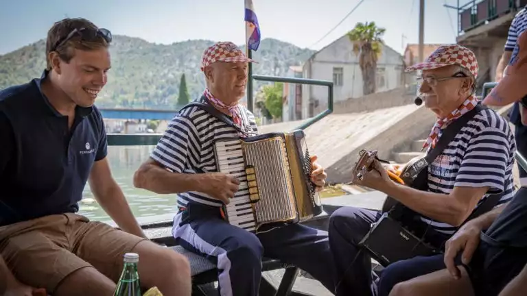 Two older men in blue and white striped shirts and checkered red hats play accordion and guitar with a younger man in a blue shirt and khaki shorts smiles beside them