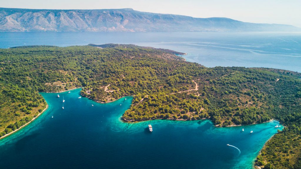 A small yacht is seen way down by the shore, among other small boats, on a remote shoreline in Croatia