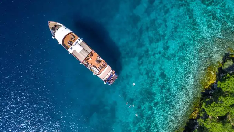 Aerial view of Aurora Croatia yacht seen near a green treed shoreline with turquoise water and five swimmers in the water