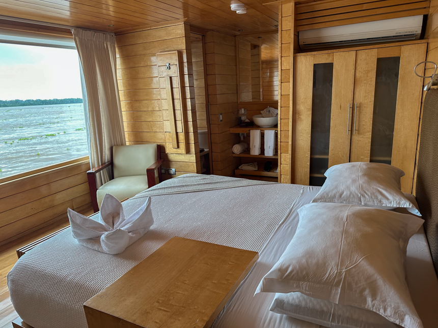 A white queen bed looks out through a large picture window in a wooden cabin aboard Delfin II Peruvian amazon river boat. 