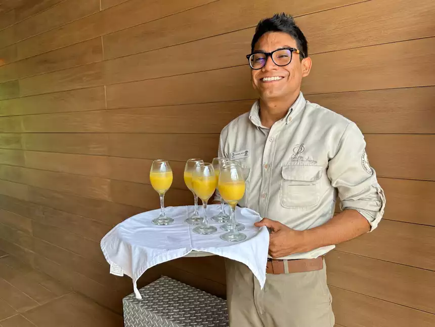A crew member from Delfin river boat wearing glasses and all tan uniform holds a white tray balancing glasses with yellow liquid. 
