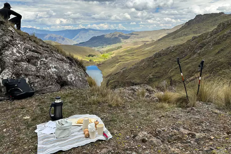 Blanket with a picnic of drinks, bread, cheese and other items set out on mountainside overlooking high alpine lake in Peru.