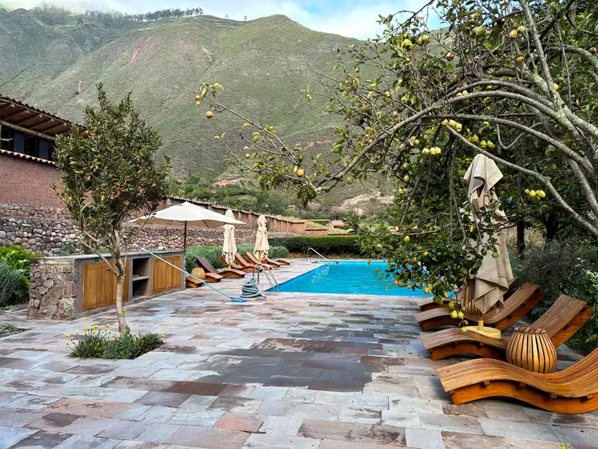 Outdoor pool area with wooden sun loungers, umbrellas line up around, surrounded by fruit trees and green mountains.