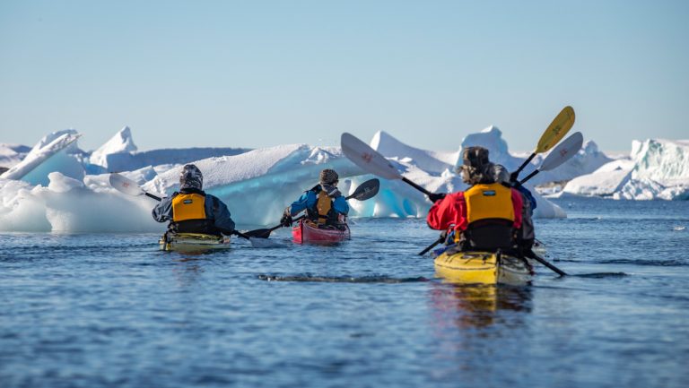 Small group of Antarctica kayakers in yellow boats paddle beside icebergs in calm water with snowcapped peaks in the distance.