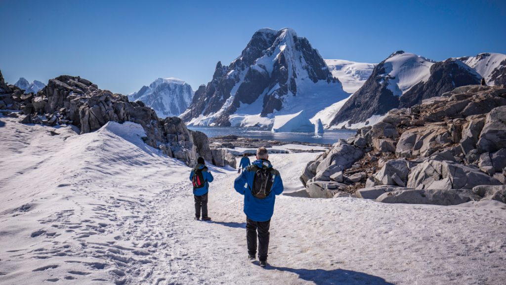 Intrepid Best of Antarctica travelers in blue parkas walk over snow beside boulders on a sunny day with snowy peaks ahead.