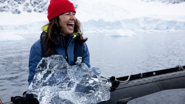 Woman in red knit hat smiles while sitting in inflatable dinghy & holding chunk of clear ice on the Intrepid Best of Antarctica.
