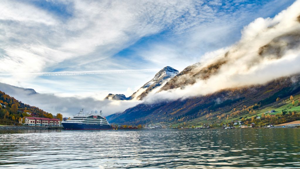 Small ship with blue hull & white upper decks sits docked in calm fjord by autumnal mountains shrouded in mist, in Norway.