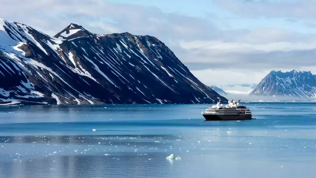 Small expedition ship with dark hull & white upper decks cruises a Norwegian fjord in winter, among iceberg bits & snowy peaks.