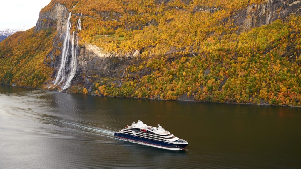 Small ship with blue hull & white upper decks cruises through calm Norwegian fjords with fall colors on hillside & waterfall.