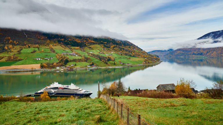 Small ship with blue hull & white upper decks sits docked along a grassy field in a glassy fjord in Norway.