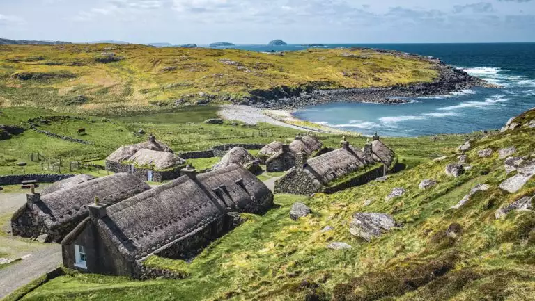 Aerial view of Hebrides, Scotland, with rugged coast, grassy hills & dark brown barn-like buildings nestled together.