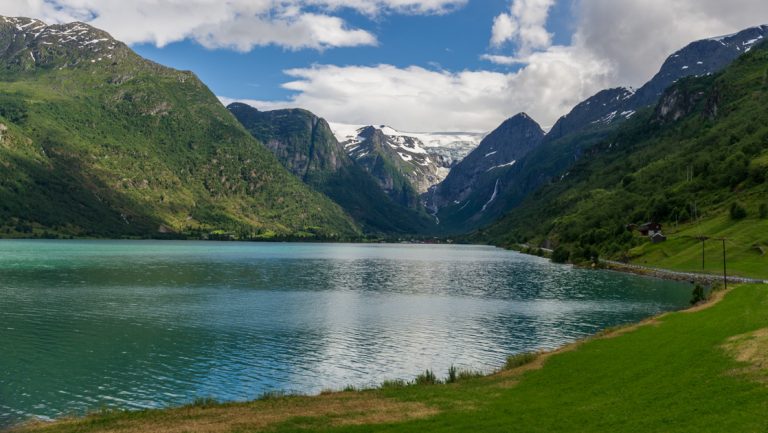 Calm fjord in Norway with teal water, bright green grass & verdant mountains under a partly cloudy sky.