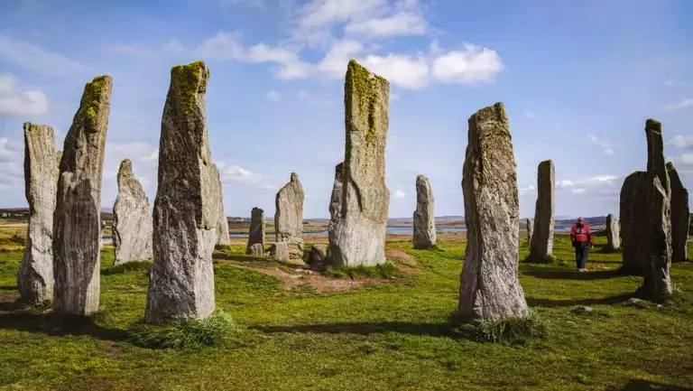 Scottish Isles & Norwegian Fjords cruise guest in red coat walks green grass among a monolith of standing stones under blue sky.