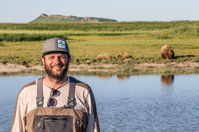 A male traveler in a brown overalls and grey AdventureSmith logo hat smiles at the camera with brown bears on the shoreline blurred in the background behind him