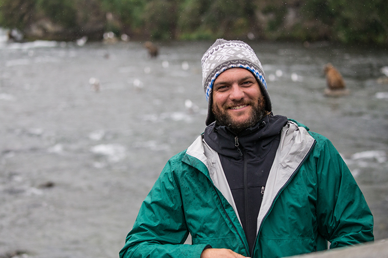 A male traveler in a green jacket and grey beanie smiles at the camera with bears in a river blurred in the background behind him