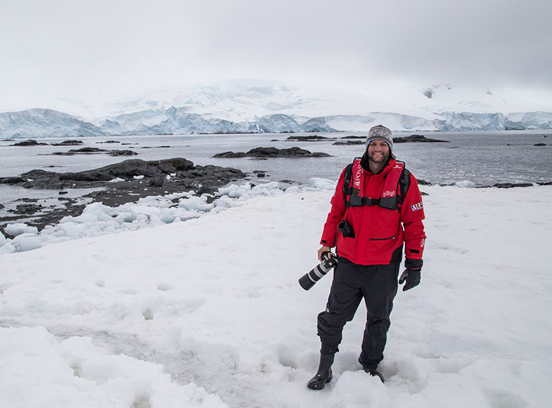 A man in a beanie and red jacket holds his camera while standing on snow at the shore in Antarctica with icy mountains behind him