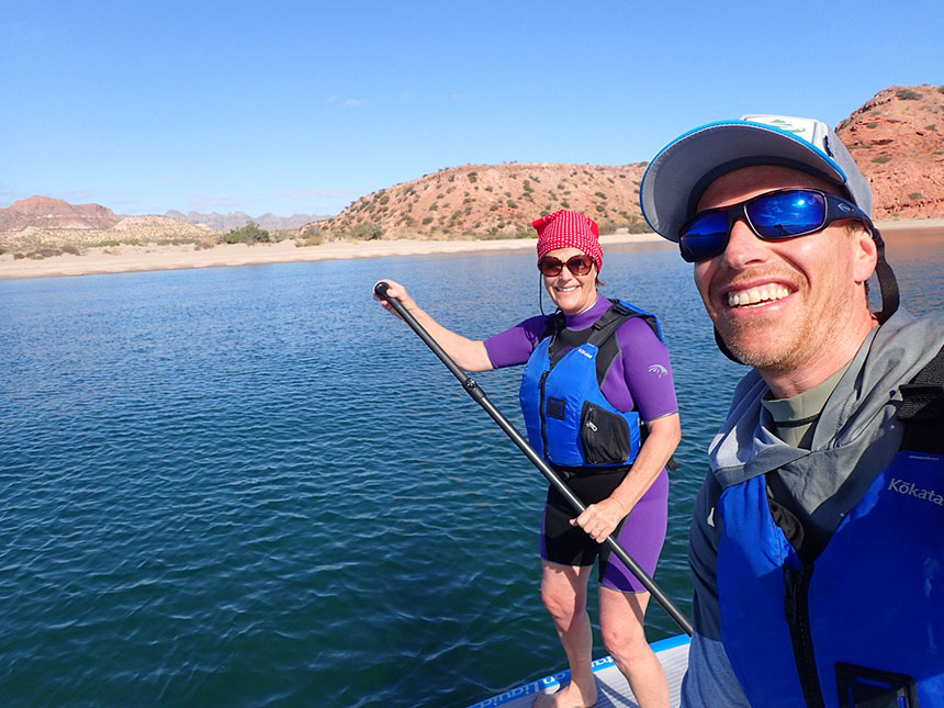 Two travelers on stand-up paddleboards in the ocean pose for a selfie in front of a deserted desert beach in Baja