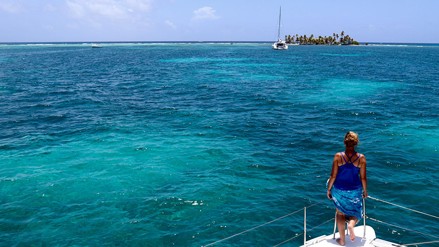 A woman seen from behind standing on the bow of a ship heading towards a Belize atoll in turquoise water