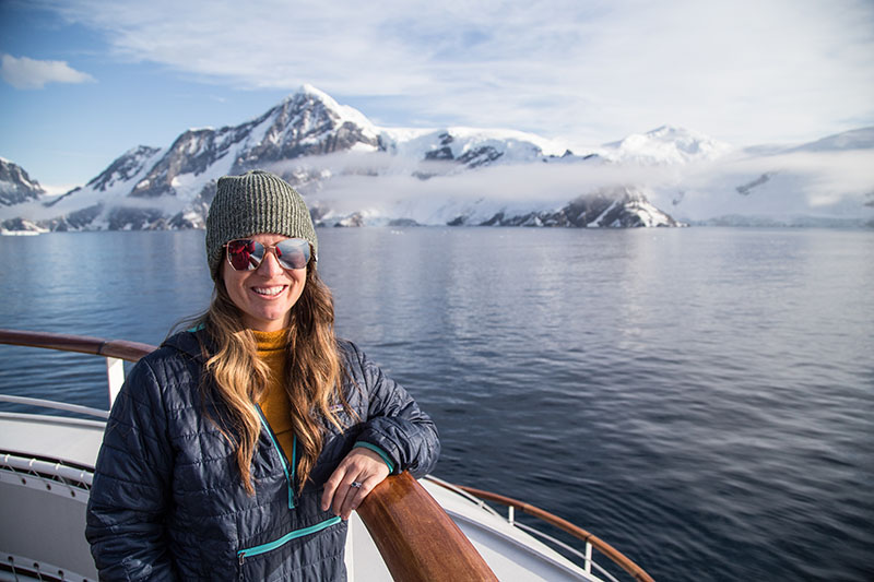 A female traveler in sunglasses and a blue jacket leans against the railing of a ship in Antarctica with the calm ocean and white-capped mountains behind her