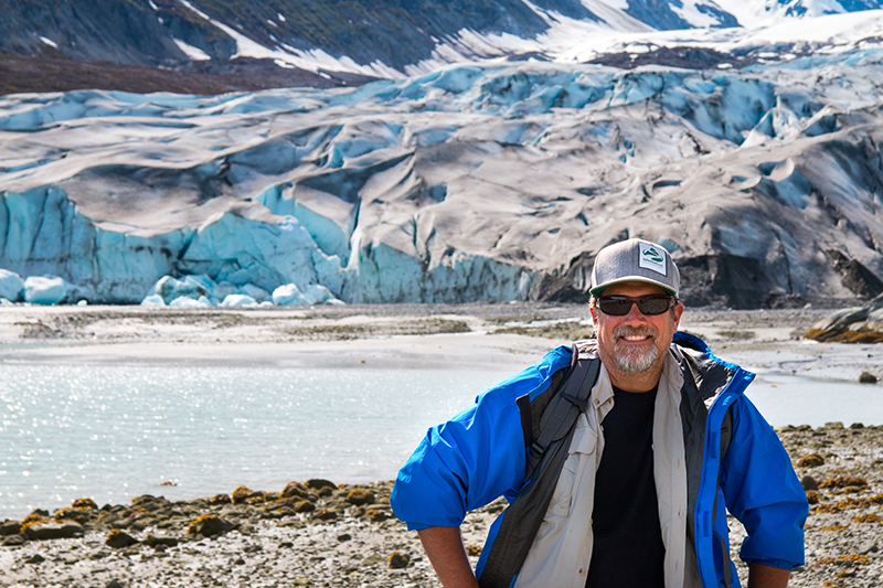 A man in a blue jacket and grey hat stands smiling on the shore with a big blue glacier behind him