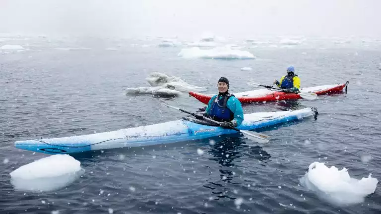A man in teal jacket paddles a blue single kayak next to another man in yellow jacket and red kayak as it snows in Antarctica