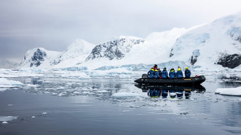 A black inflatable boat carried cruise guests in royal blue parkas and cruises through icebergs in Antarctica landscape