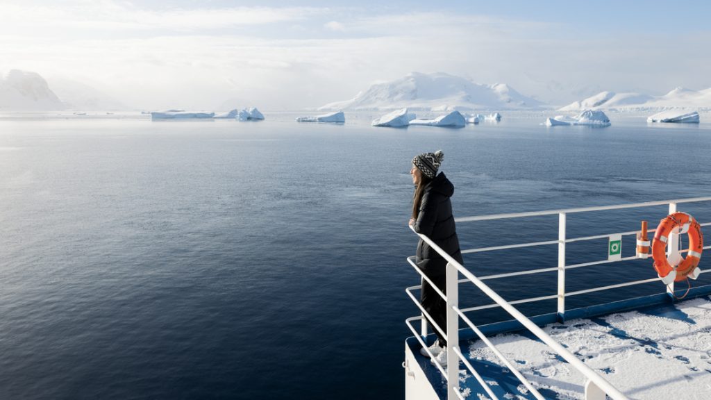 a female traveler wearing a beanie leans over white ship railing looking over an icy ocean landscape in antarctica