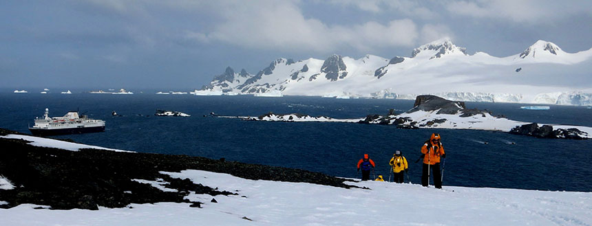 Four skiers head up a snowy hill with the Ocean Endeavour ship down in the ocean below them with big snowy mountains off to the side