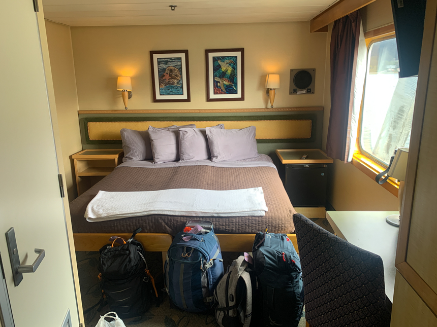 Admiral category stateroom #201 on the Cabin Deck of Safari Endeavour. Yellow walls, window two lamps, art, and a queen bed, 
