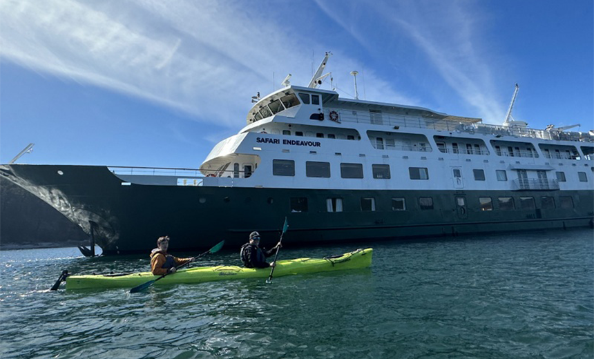 In Alaska two men paddle a lime double kayak in front of the white and green Safari Endeavour small ship. 