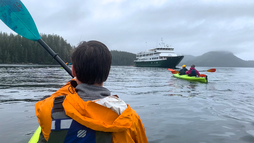 On an overcast grey day in Alaska kayakers paddle double kayaks towards the white and green Safari Endeavour ship. 