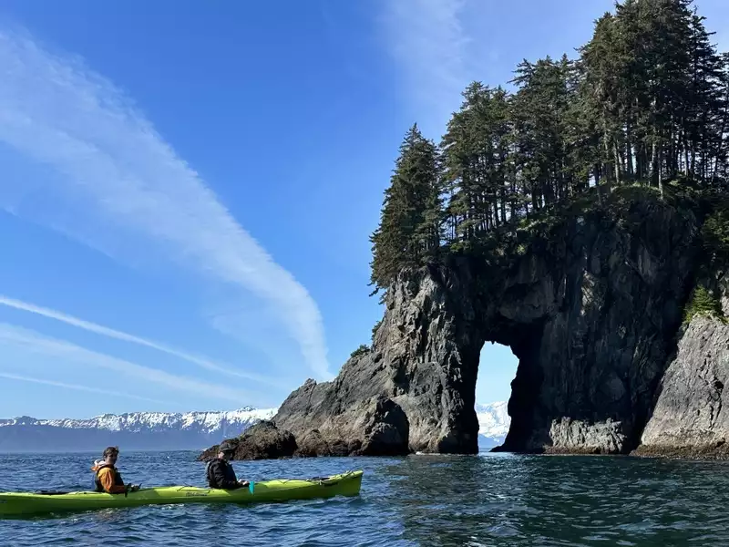 Two men kayaking a lime double kayak in front of a magnificent rock arch with whitecapped peaks in the background