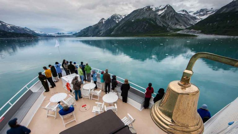 Travelers stand on bow of small ship with gold bell & white tables looking onto teal water with snowcapped peaks behind.