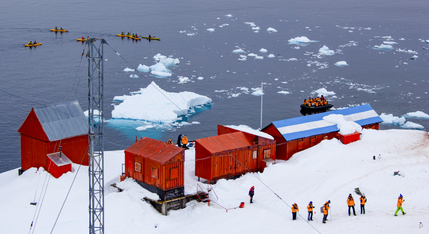 December in Antarctica, guests are able to visit polar research stations. Multiple red buildings along shoreline on white snow