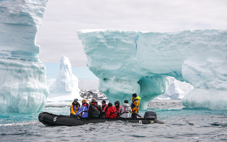 on an overcast day guests ride a black inflatable skiff around massive icebergs floating in Antarctica.