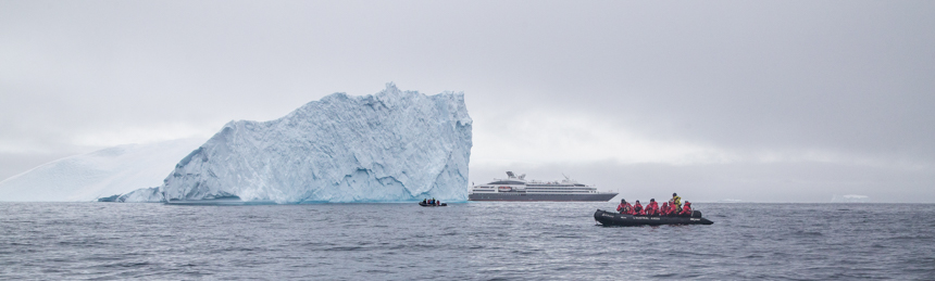 Photo compares the an iceberg the size of a building to a 200 person ship to a small inflatable skiff carrying pasengers in red parkas. 