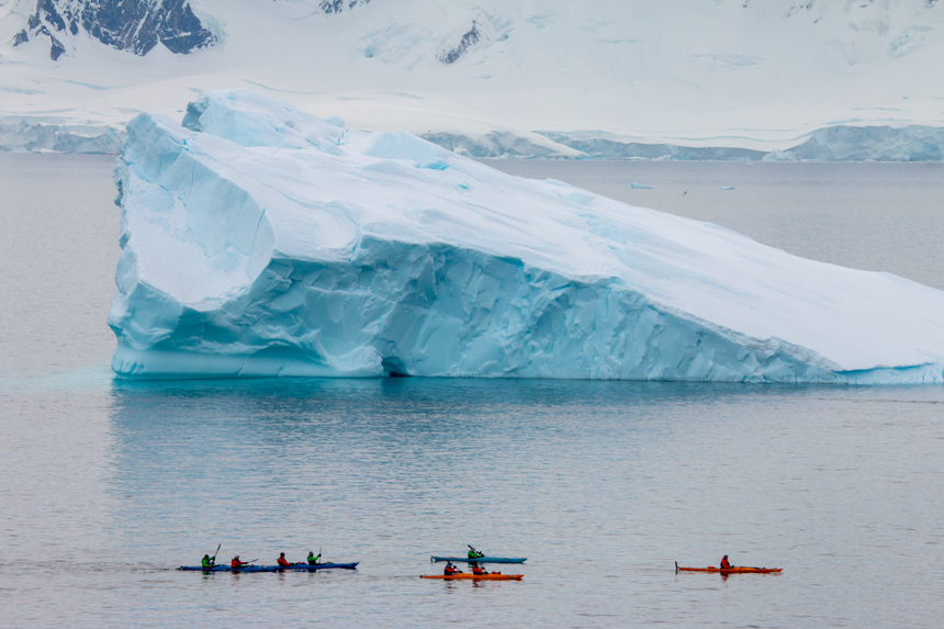 A group of kayakers in blue green and red kayaks paddle around a massive icy blue and white iceberg floating in the Antarctic ocean. 