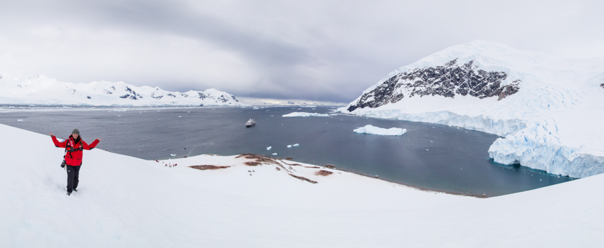 Girl in red parka stands on top of snow covered hill overlooking a blue bay with an expedition ship floating in Antarctica 