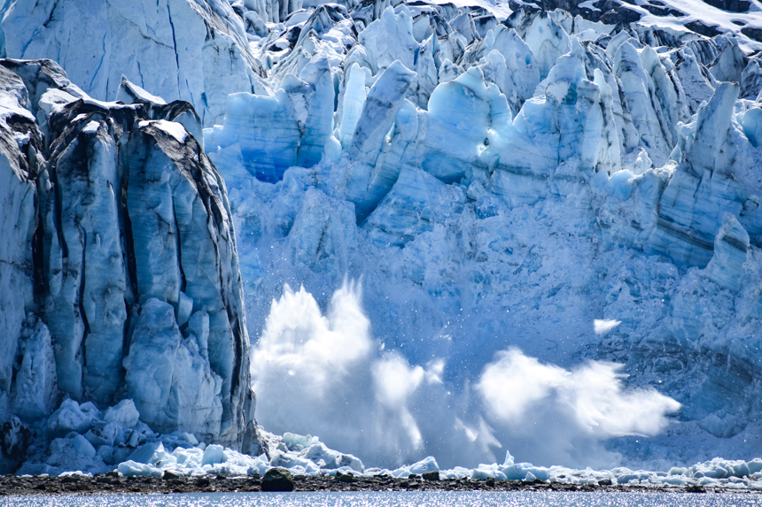 A giant splash below a massive jagged teal blue and white glacier from a calving event where ice breaks off and falls into water.