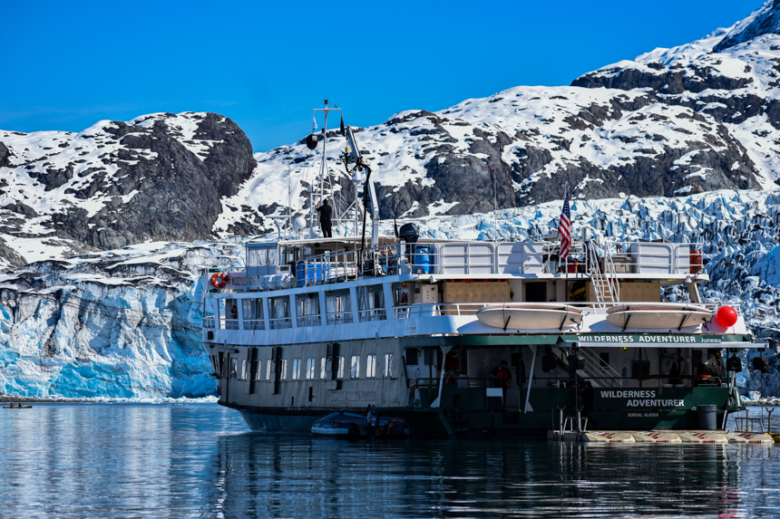 The white and green small ship Wilderness Adventure floats closely in front of a massive icy glacier during a Glacier bay Alaska cruise.