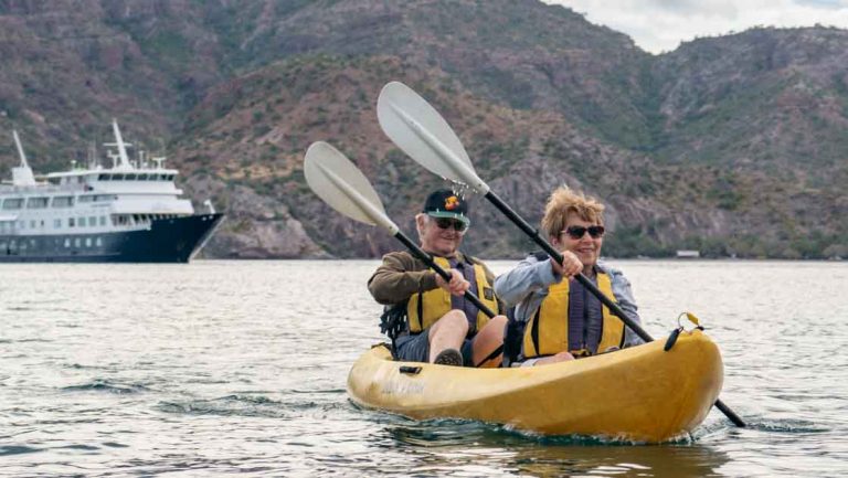 Man & woman paddle a yellow tandem kayak on calm water with a small blue & white ship & rocky hillside behind in Baja.