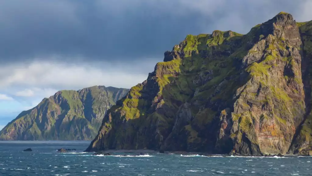 Jagged volcanic mountains of rock with green moss rise from dark blue seas in the Aleutian Islands, Alaska. on a cloudy day.