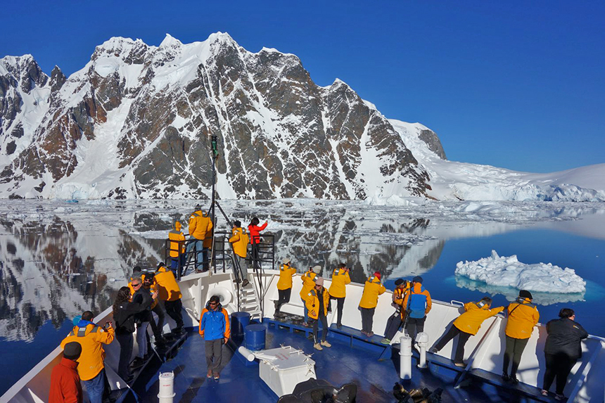 Guests in yellow parkas gather at bow of ship for views of snow covered mountains in Antarctica.