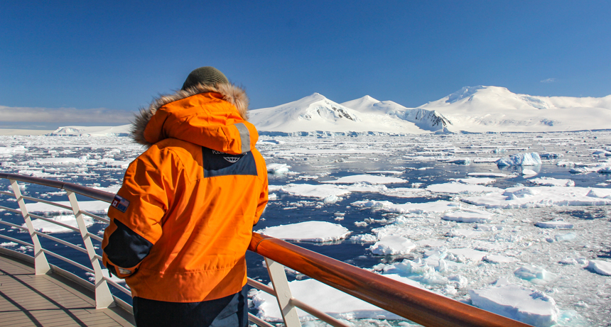 A man in orange parka leans on railing and looks out over an ice filled ocean landscape in March in Antarctica,