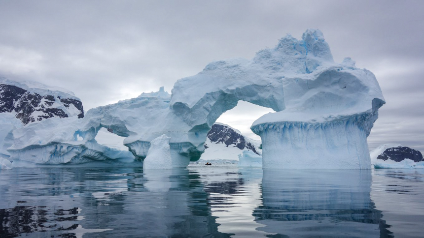 An overcast grey day in Antarctica, a teal iceberg with multiple arches melted from the suns rays floats in the water. 