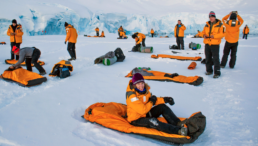 A group of Antarctica cruise travelers wearing yellow parkas set out their orange sleeping bags on the white snow for a camping activity. 