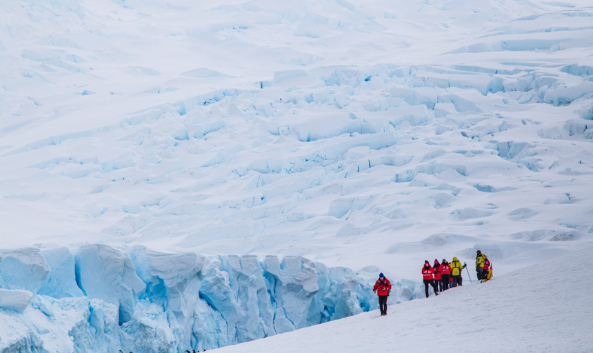 Guests in red parkas stand on snowy hillside surrounded by while snow covered glaciers in Antarctica in October 