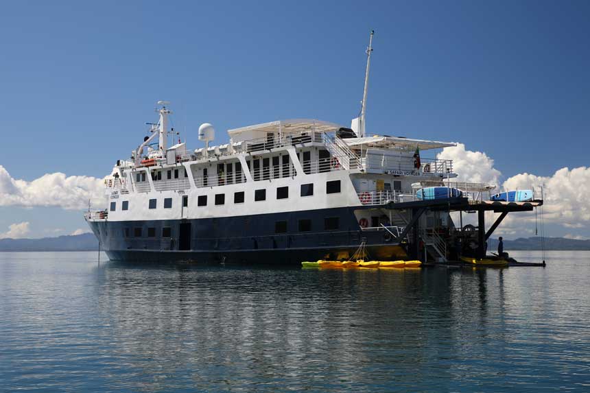 Small ship with dark blue hull & 3 white upper decks sits in calm water on a sunny day with yellow kayaks sitting empty beside.