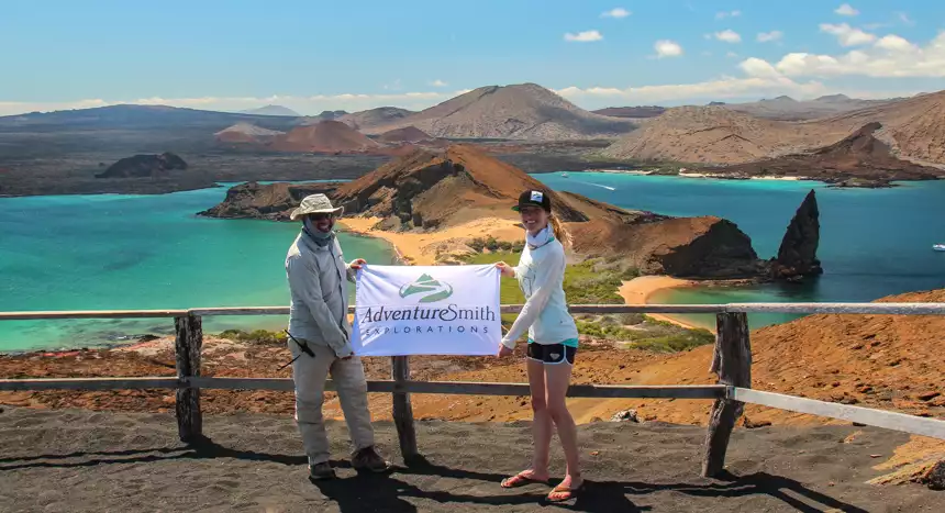 Male and female hold AdventureSmith Explorations logo flag at the top of a vista point overlooking beautiful Galapagos island volcanic landscape. 