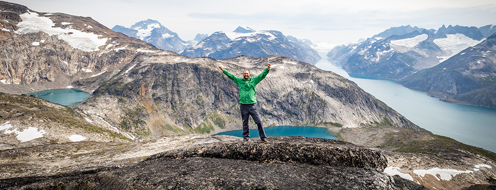A male traveler in a green jacket standing with arms raised on a mountain top overlooking green lakes and a blue fjord with snowcapped mountains in the arctic. 